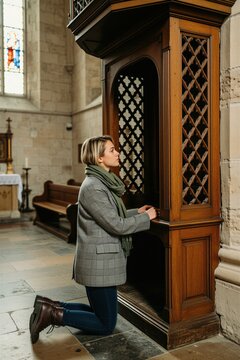 Young woman kneels in confession booth inside church. She wears grey checkered jacket and green scarf. Female seeks spiritual guidance praying. Confessionals are traditional part of church worship.