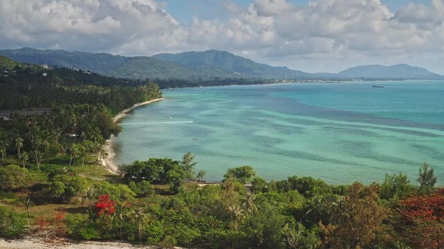 Amazing Koh Samui lush tropical coastline stretches along tranquil white sand beach. Drone flight over crystal turquoise bay, green palm trees, mountains in the background. Thailand travel destination