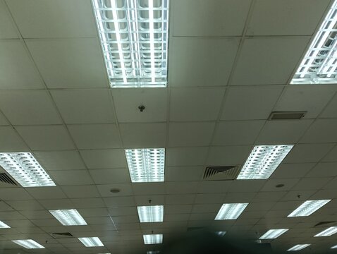  view of a commercial office ceiling featuring multiple recessed fluorescent LED light fixtures, suspended ceiling tiles, air conditioning ventilation grilles, and mounted display monitors. The interi