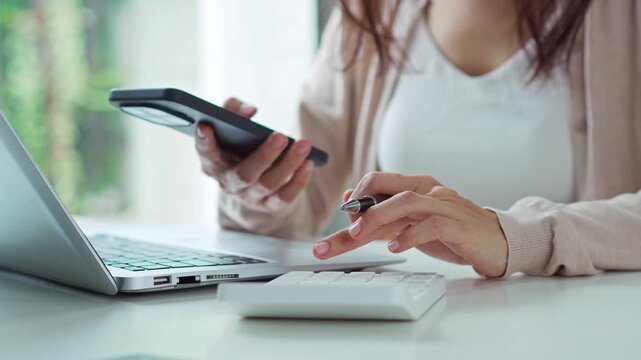 Pensive young Asian woman in home office calculating monthly household expenses and utility bills using a calculator