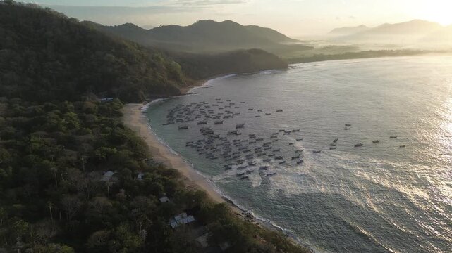 Aerial view of Papuma Beach with numerous small fishing boats anchored in the bay, surrounded by lush green hills and sandy coastline in East Java, Indonesia.