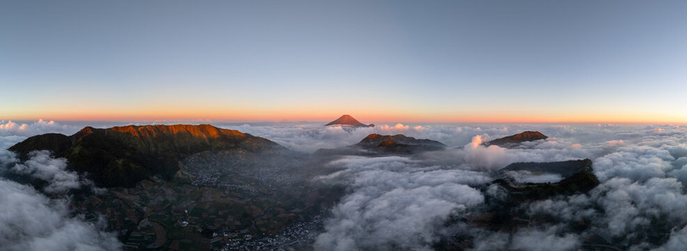Aerial view of Dieng Plateau at sunset, with volcanic peaks rising above a dense sea of clouds and terraced agricultural fields in the valley below, Kejajar, Central Java, Indonesia.