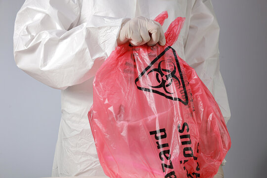 Cautious worker wearing protective hazmat suit with glove holds red biohazard bag filled with toxic waste during safe medical disposal