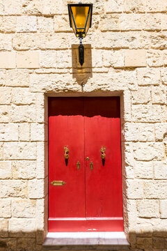 Mdina, Malta: A vivid red door set in a limestone wall, featuring ornate brass knockers, a lantern above the entrance and warm Mediterranean light highlighting the historic architecture of the town