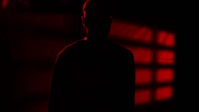 Mysterious male silhouette emerging from dark red-lit room, reaching out and leaving bloody handprint on glass surface with dramatic shadow pattern. Cinematic thriller and horror concept for crime, my