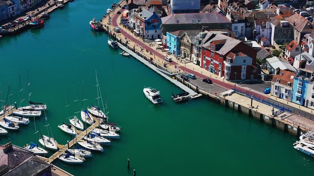 Drone view over sailing boats moored beside the colourful waterfront and quay at Weymouth Harbour in Dorset on a sunny summer day.