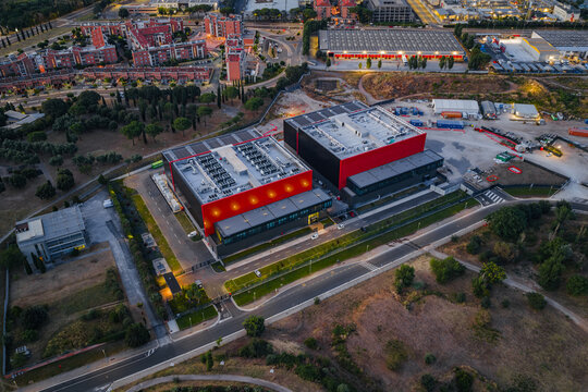 Aerial view of a modern datacenter facility featuring red and black exterior walls and rooftop cooling systems, situated near roads and urban infrastructure in Settecamini, Lazio, Italy.