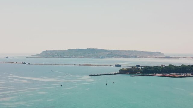 High drone view across Weymouth Bay, showing calm turquoise water, harbour walls, anchored boats, Nothe Fort and the Isle of Portland in the distance.