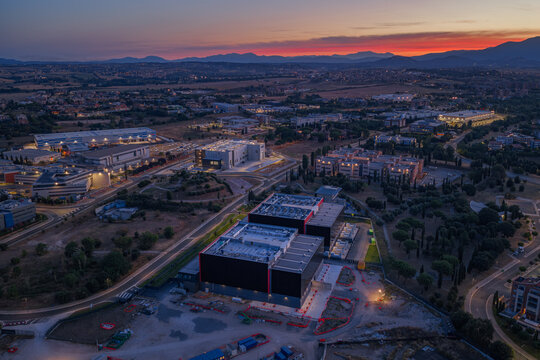 Aerial view of a modern datacenter complex at sunset, featuring large industrial buildings, surrounding roads, and trees in Settecamini, Lazio, Italy.