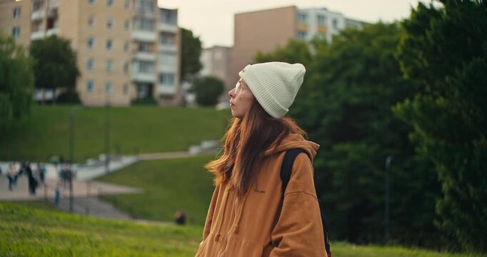Portrait of a woman wearing a beanie and backpack in a green urban park. Soft natural light and a blurred city background create a relaxed outdoor lifestyle scene.