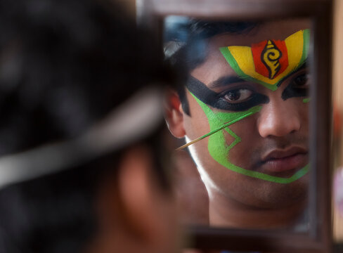 Kathakali dancer looking in the mirror and applying green colour on his face. 