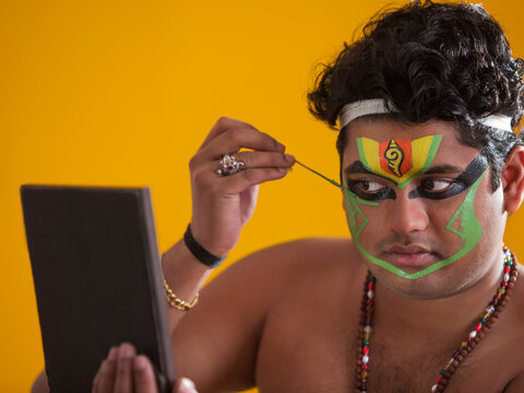 Kathakali dancer looking in the mirror and applying green colour on his face. 
