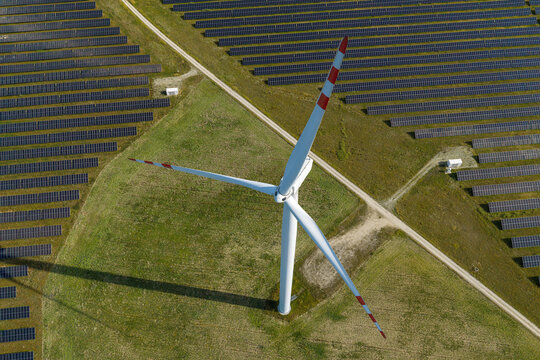 Aerial view of a large wind turbine standing in the middle of a vast solar farm with rows of photovoltaic panels in a green field in Poland.
