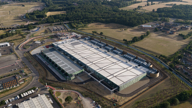Aerial view of a large industrial data center facility with green facades and rooftop cooling units, surrounded by access roads and green fields in Waltham Cross, England, United Kingdom.