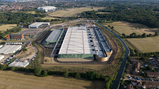 Aerial view of a large modern data center facility with solar panels on the roof, surrounded by green fields, a canal, and suburban housing in Waltham Cross, England, United Kingdom.