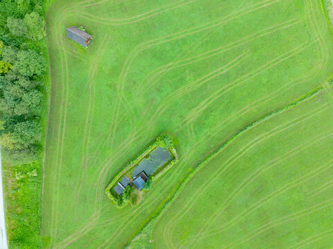 Aerial view of Nationalpark Gesause featuring a small pond surrounded by lush green meadows, rustic wooden structures, and intricate tire tracks in Admont, Steiermark, Austria.