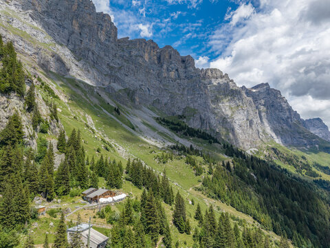 Aerial view of the Tallihutte SAC mountain hut nestled among steep rocky cliffs, lush green meadows, and dense coniferous forests in Innertkirchen, Bern, Switzerland.