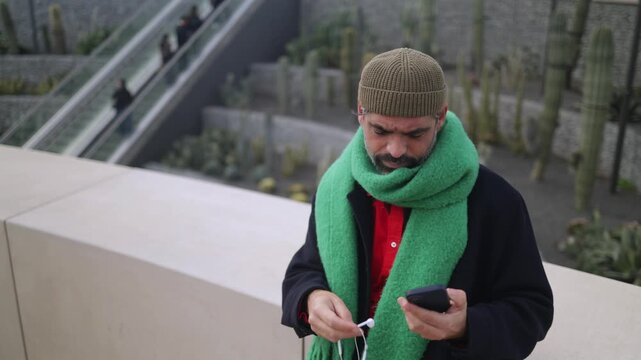 Mature man untangling wired earphones for listening music on his smartphone in urban cactus park, showcasing modern life struggles and connectivity