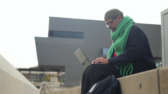 Focused man with a beard and glasses working as a freelancer on his laptop, sitting on concrete steps in a modern urban environment, embodying the digital nomad lifestyle