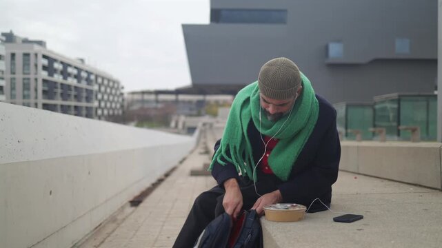 Mature man with a green scarf listening to music with earphones and preparing for a lunch break in an urban setting, enjoying a takeaway meal with a reusable bottle