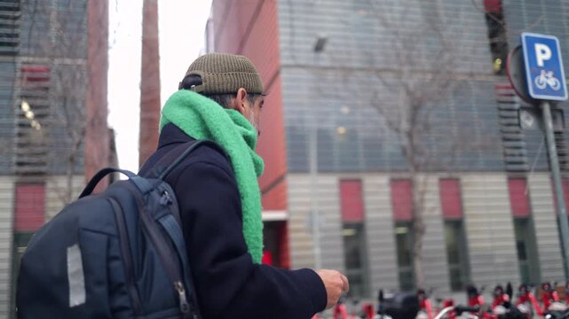 Mature man with a green scarf and backpack walking on a modern city street in winter. Urban pedestrian commuting with a modern building and bike sharing station in the background