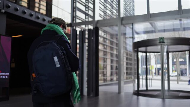 Young man with a backpack walking through a modern business center lobby, passing through an electronic turnstile gate and heading towards a revolving door to the street