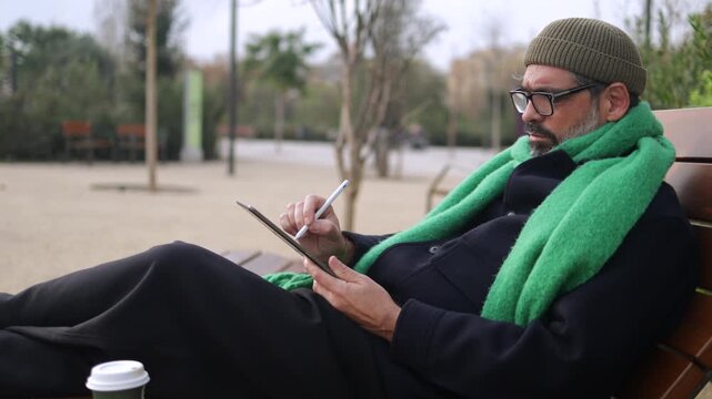 Mature man relaxing on a park bench and using a digital tablet with a stylus, creative professional working remotely in a public space on a chilly day with a cup of coffee