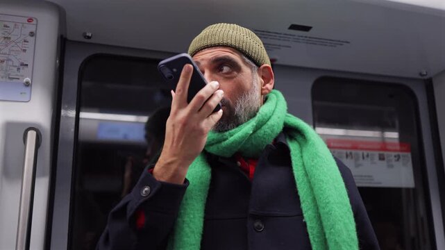 Mature bearded man in a beanie and green scarf using a smartphone to send a voice message while commuting on a public subway train, staying connected on the go