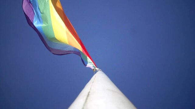 Stockholm, Sweden A Pride flag flies on a flagpole against a blue sky. 