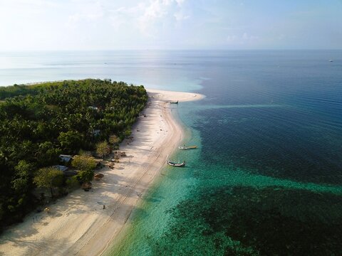 Aerial view of a tropical beach with turquoise water, white sand, lush palm trees, and traditional boats moored along the shore in Sumenep, East Java, Indonesia.