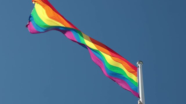 Stockholm, Sweden A Pride flag flies on a flagpole against a blue sky. 