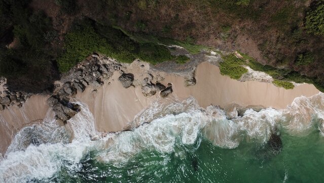 Aerial view of a secluded beach with rocky cliffs, white sand, and turquoise ocean waves crashing against the shore in Indonesia.