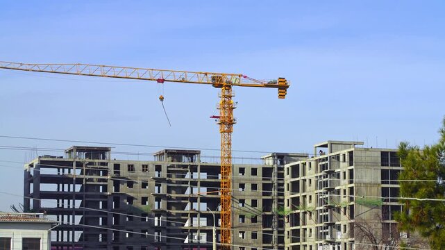 Video footage from a construction site where a yellow tower crane is being used to erect a multi-story building. The raised hook and free slings swing, and the boom swings toward the loading area