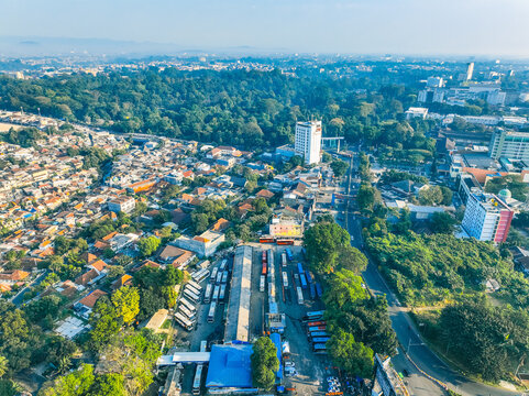Aerial view of the Baranangsiang bus terminal featuring rows of parked buses, surrounding urban residential areas, dense green trees, and city buildings in Bogor Timur, West Java, Indonesia.