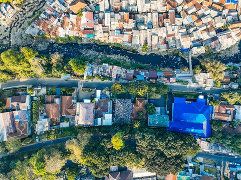 Aerial view of the Baranangsiang district featuring dense residential housing, a winding river, lush trees, and a prominent blue-roofed building in Bogor Timur, West Java, Indonesia.