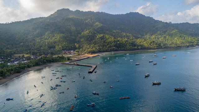 Aerial view of Pantai Karanggongso featuring numerous fishing boats anchored near a concrete pier, surrounded by lush green mountains and calm blue waters in Watulimo, East Java, Indonesia.