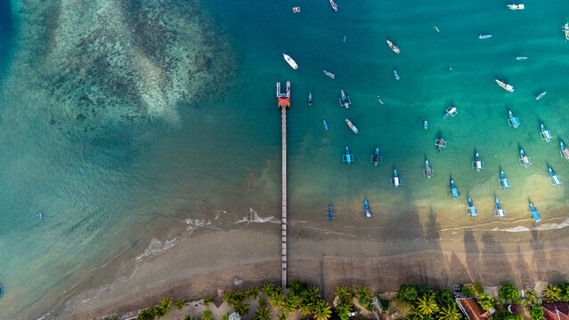 Aerial view of Pantai Karanggongso featuring a wooden pier extending into the turquoise bay filled with fishing boats, with a sandy beach and palm trees in Watulimo, East Java, Indonesia.