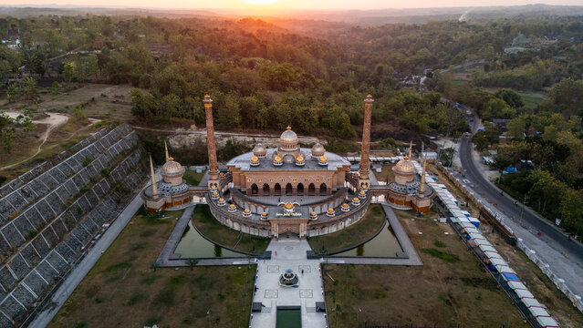Aerial view of the ornate Masjid Al-Ba'its with its multiple domes and tall minarets surrounded by lush greenery and reflecting pools during a golden sunset in Bojonegoro, East Java, Indonesia.