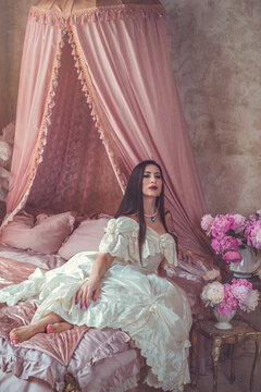 Sensual dark-haired lady posing on a velvet bed surrounded by blooming pink peonies, luxury fragrance and beauty campaign concept in a vintage boudoir setting