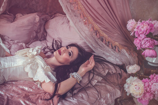Sensual dark-haired lady posing on a velvet bed surrounded by blooming pink peonies, luxury fragrance and beauty campaign concept in a vintage boudoir setting