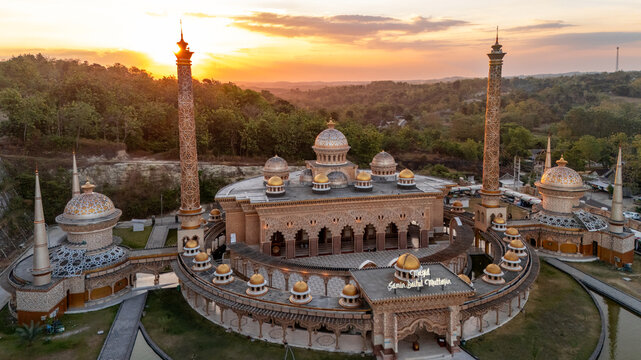 Aerial view of Masjid Samin Baabul Muttaqien with its intricate golden domes and tall minarets surrounded by lush greenery during a warm sunset in Bojonegoro, East Java, Indonesia.