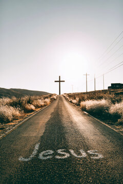A straight road leads toward a silhouette of a large cross with the word Jesus written on the ground. Dry fields surround the road under warm light