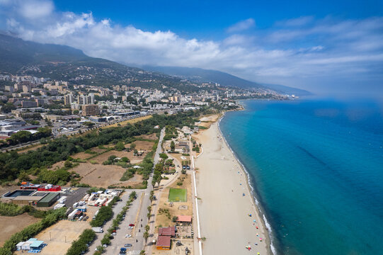 Aerial view of L'Arinella beach with its sandy coastline, turquoise Mediterranean sea, and the urban landscape of Bastia under a blue sky in Bastia, Corse, France.