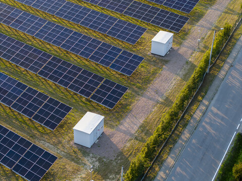 Aerial view of rows of solar panels and white electrical cabinets in a field with green grass and a nearby road in Krakow, Lesser Poland Voivode, Poland.
