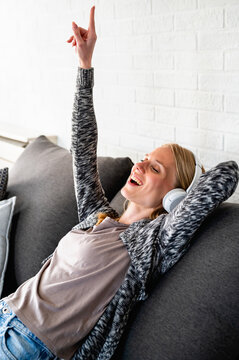 Happy young woman listening to music with wireless headphones while relaxing on a sofa at home. Expressing joy, freedom and positive emotions in a cozy modern living room