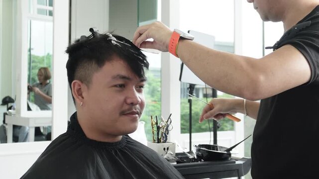 Hair stylist sectioning hair and placing foils for highlights during professional hair highlighting treatment for an Southeast Asian man in his early 20s