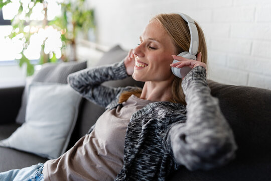 Happy young woman relaxing on a cozy sofa at home, listening to music with wireless headphones. Peaceful moment of self-care, stress relief, comfort and mindfulness in a modern living room.