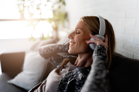 Happy young woman relaxing on a cozy sofa at home, listening to music with wireless headphones. Peaceful moment of self-care, stress relief, comfort and mindfulness in a modern living room.