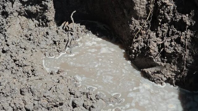 Macro view of bubbling muddy water in a street trench, highlighting environmental pollution and resource loss from a ruptured underground pipe.