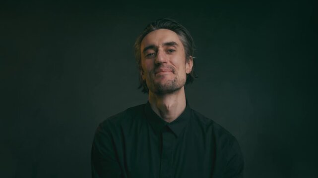 Calm young adult man with closed eyes and slight smile sitting against dark blurred background in low-key lighting